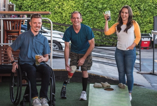 Two men and one woman playing cornhole. One man is in wheelchair and the other man has a prosthetic leg.