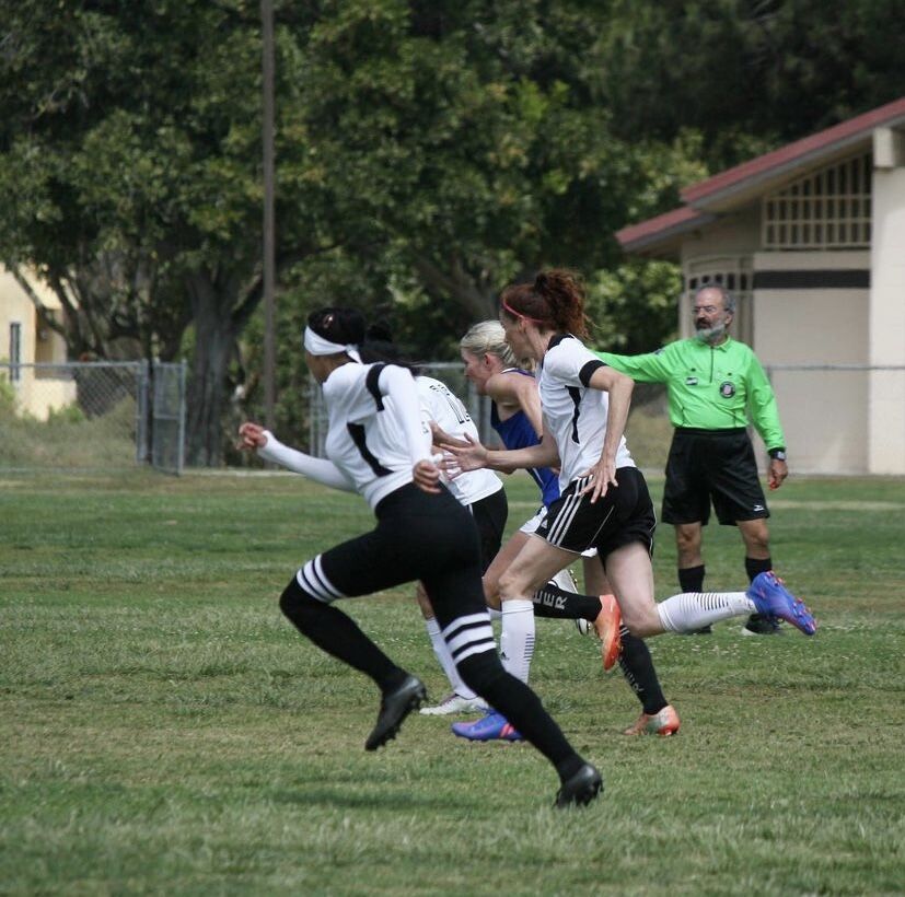 Three women in white jerseys and one in blue running down the soccer field.