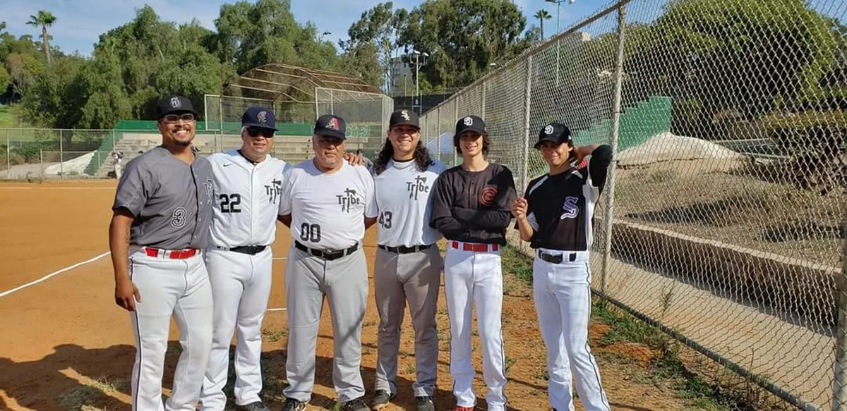 Three generations of Carrillo men in baseball uniforms posing together on baseball field.