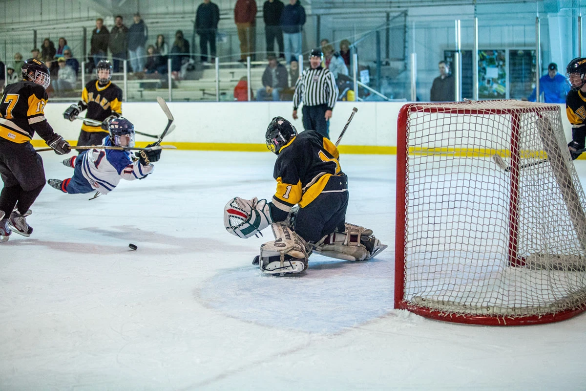 Hockey players in a game with one player diving in the air toward the goalie as goalies teammates are trying to stop him from scoring.
