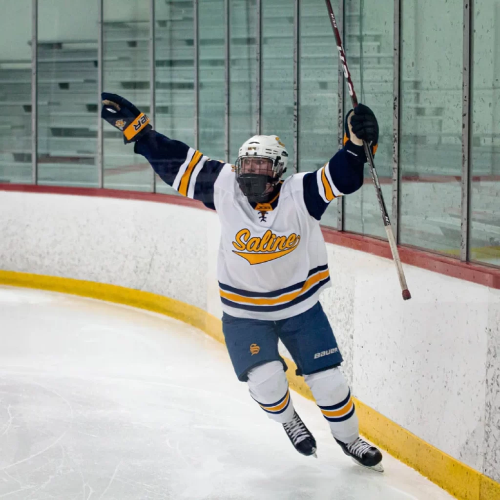 Hockey player on the ice with both hands raised in air holding hockey stick up in one hand.