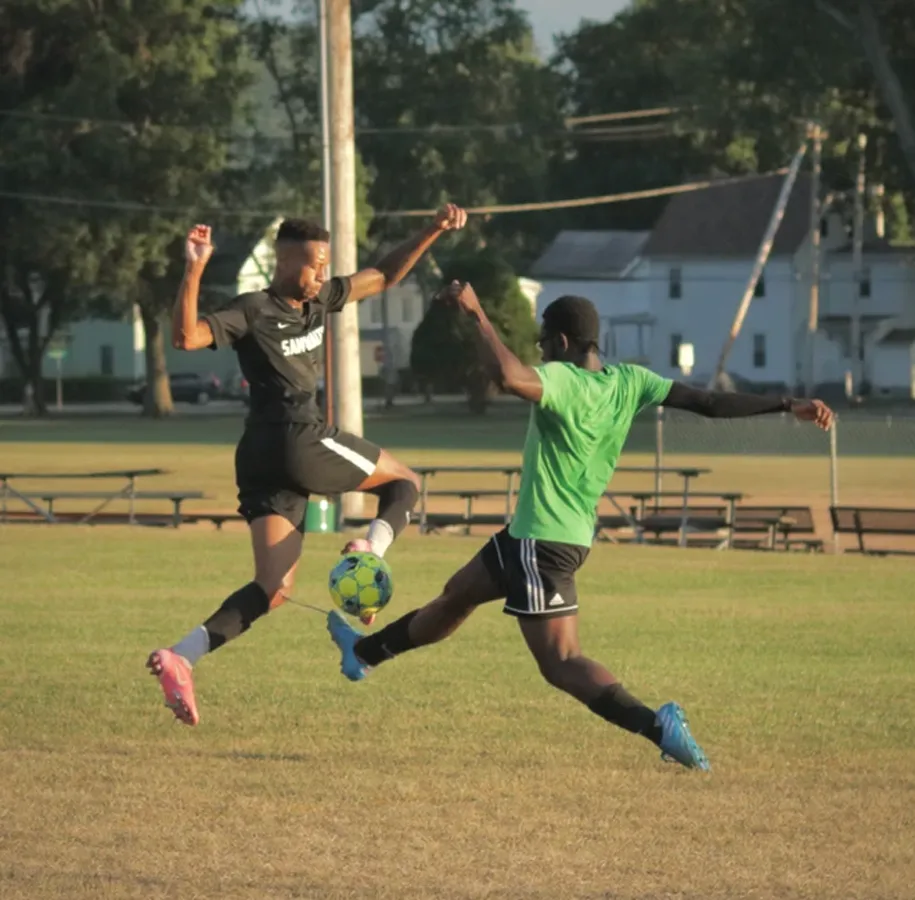 Two male adult soccer opponents on field both in air with legs out, arms outstretchd, and ball in between them